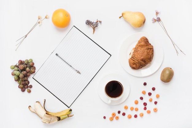 Stift auf einem offenen Notizbuch mit Kaffeetasse; Croissant und viele Früchte auf weißem Hintergrund