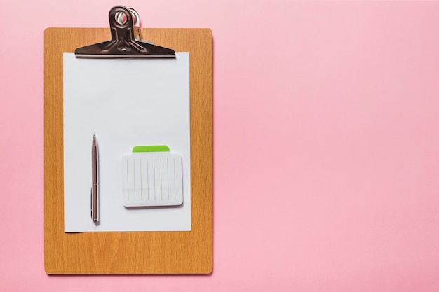 Stift und Notizblock auf leerem Papier über hölzernem Klemmbrett gegen rosa Hintergrund