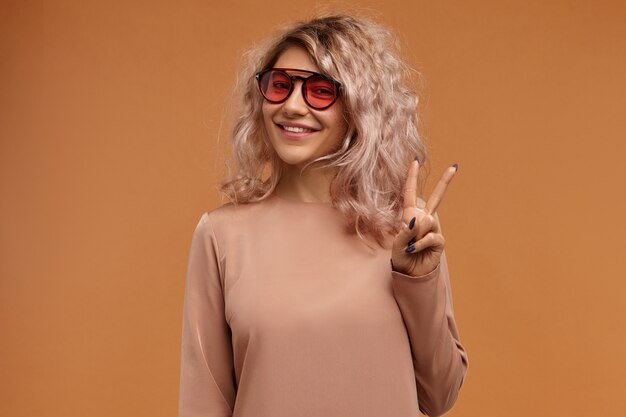 Stil, Modetrends und Jugendkonzept. Bild des modischen Hipster-Mädchens, das trendige Sonnenbrille mit rosa Gläsern mit freudigem breitem Lächeln trägt, Friedenszeichen machend