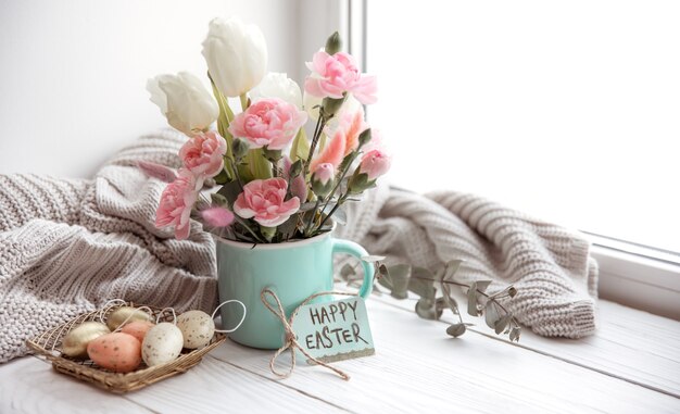 Stillleben mit frischen Frühlingsblumen in einer Vase, Eiern, einer Frohe Osterkarte und einem gestrickten Element.