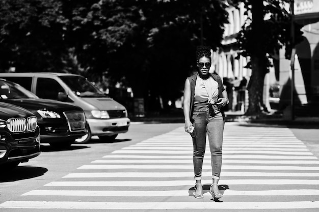 Stilvolle afrikanisch-amerikanische Frau, die mit dem Handy zur Hand auf dem Zebrastreifen oder Fußgängerüberweg geht