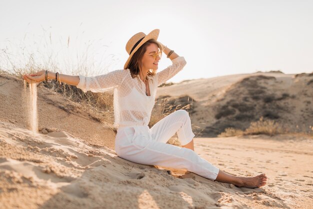 Stilvolle attraktive lächelnde Frau, die im Wüstensand posiert, gekleidet im weißen Kleidungsoutfit, das Strohhut und Sonnenbrille auf Sonnenuntergang trägt