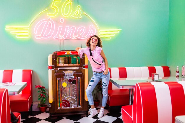 Stilvolle attraktive lächelnde Frau im Retro-Vintage-Café der 50er Jahre in der Musik-Jukebox, die Milchshake-Cocktail im Hipster-Outfit trinkt und Spaß beim Lachen in fröhlicher Stimmung hat