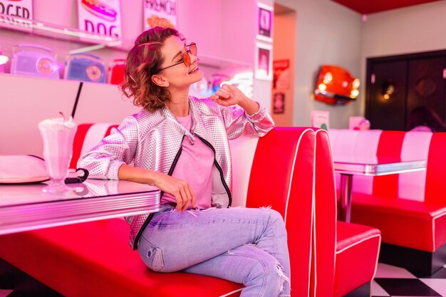 Stilvolle attraktive lächelnde Frau im Retro-Vintage-Café der 50er Jahre in rosa Farbe, die am Tisch sitzt und Milchshake-Cocktail im Hipster-Outfit trinkt und Spaß hat