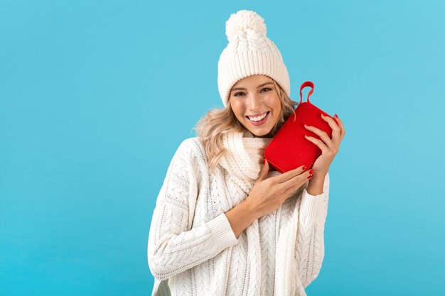 Stilvolle blonde lächelnde schöne junge Frau, die drahtlosen Lautsprecher hält, der Musik hört, die glücklich weißen Pullover und Strickmütze trägt, die auf Blau aufwirft