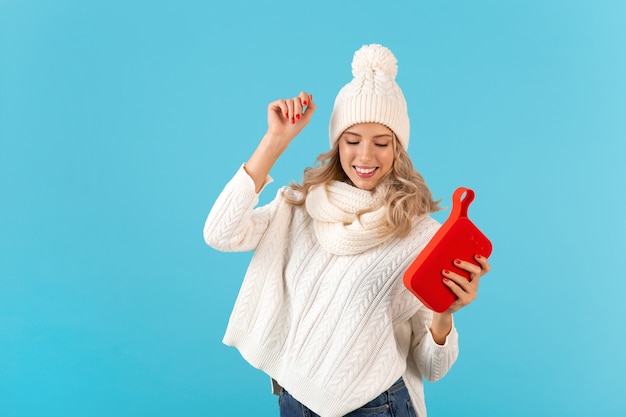 Stilvolle blonde lächelnde schöne junge Frau, die einen drahtlosen Lautsprecher hält, der Musik hört, die glückliches Tanzen mit weißem Pullover und Strickmütze im Winterstil-Mode posiert