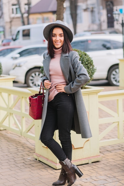 Stilvolle charmante junge Frau im grauen Mantel, Hut mit roter Tasche, die auf Straße im Stadtzentrum geht. Brünettes Haar, elegante Frau, modisches Modell, lächelnd, fröhliche Stimmung.
