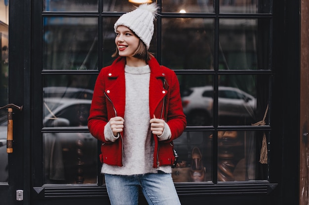 Stilvolle Dame in guter Stimmung lehnte sich an Fenster mit schwarzem Holzrahmen. Mädchen gekleidet in Jeans, Hut und rote Jacke posierend.