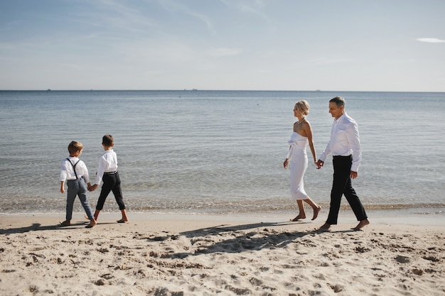 Stilvolle Familie geht am Strand in der Nähe des ruhigen Meeres spazieren, Eltern und Kinder halten sich an den Händen