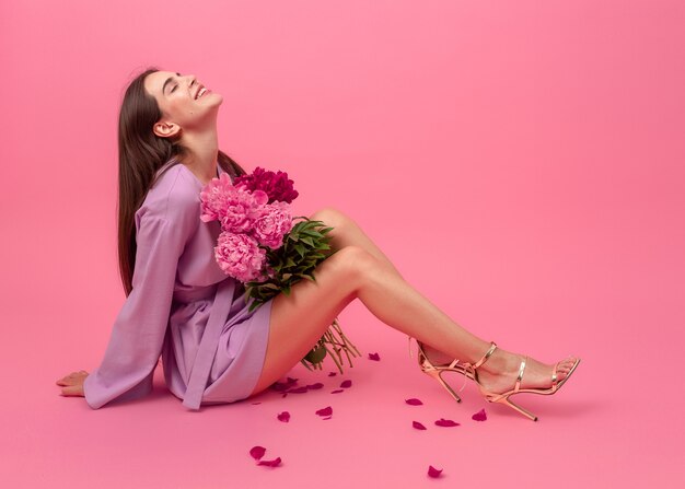 Stilvolle Frau auf rosa im Sommerviolett-Mini-Trendkleid, das mit Pfingstrosenblumenstrauß auf dem Boden sitzend aufwirft