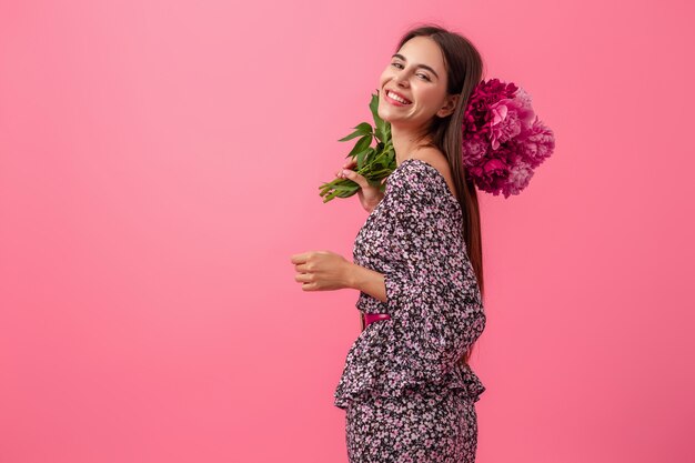 Stilvolle Frau auf Rosa im trendigen Sommerkleid, das mit Pfingstrosenblumenstrauß aufwirft