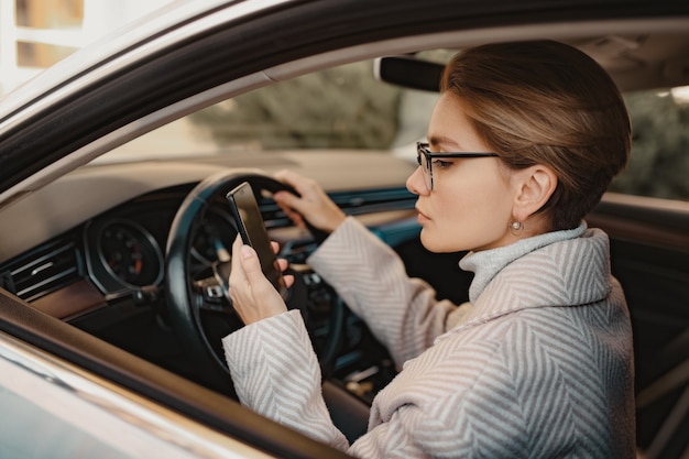 Stilvolle Frau, die im Auto sitzt, gekleidet in Mantelwinterstil und Brille mit Smartphone, Geschäftsfrau, die mit dem Fahren beschäftigt ist