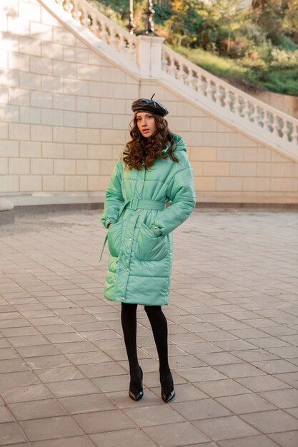 Stilvolle Frau, die im Winterherbstmode-Trend blauen Puffermantel und Hutmütze in der alten schönen Straße voller Höhe aufwirft