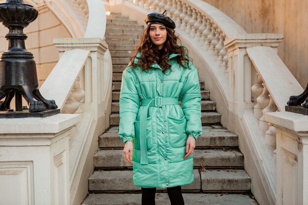 Stilvolle Frau, die im Winterherbstmode-Trend blauen Puffermantel und Hutmütze in der alten schönen Straßentreppe aufwirft