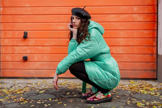 Stilvolle Frau, die im Winterherbstmode-Trendpuffermantel und in der Hutmütze gegen orange Wand in der Straße aufwirft, die bunte bedruckte Schuhe trägt