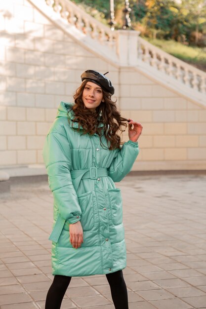 Stilvolle Frau, die im Winterherbstmodetrend blauen Puffermantel und Hutmütze in der alten schönen Straße aufwirft