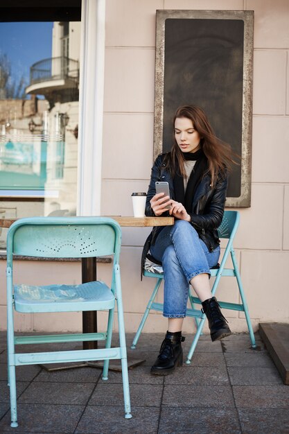 stilvolle Frau, die in der Terrasse des Restaurants sitzt, Kaffee trinkt und Termin zur Kosmetikerin über Internet, Smartphone hält und tippt