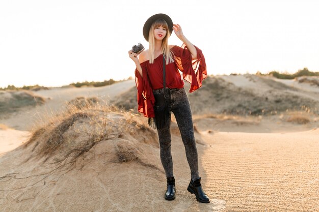 Stilvolle Frau, die in der Wüste aufwirft, schwarzen Hut und Boho-Outfit tragend