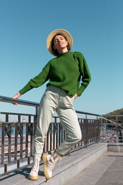 Stilvolle Frau im grünen lässigen Pullover und Hut im Freien auf Brücke mit Flussblick genießt sommerlichen sonnigen Tag