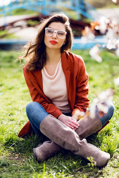 Stilvolle Frau im lässigen Frühlingsoutfit, das auf Gras im sonnigen warmen Park sitzt