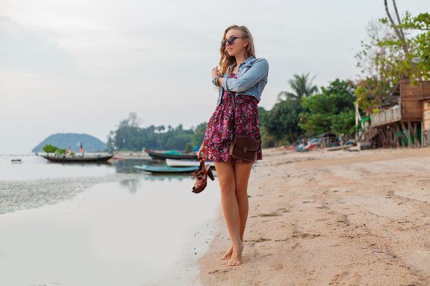 Stilvolle Frau im Sommerkleidurlaub, der am Strand mit Schuhen in der Hand geht