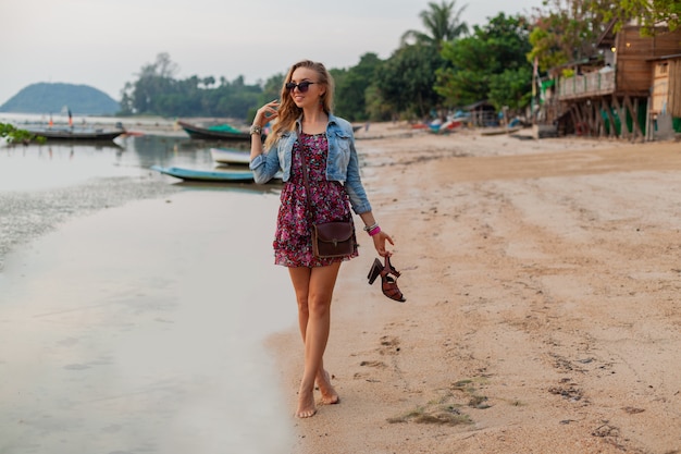 Stilvolle Frau im Sommerkleidurlaub, der am Strand mit Schuhen in der Hand geht