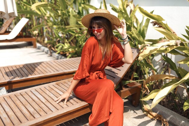 Stilvolle Frau im Strohhut und im orangefarbenen Spielanzug, die während des Urlaubs auf Bali auf ihrer Villa ruht.