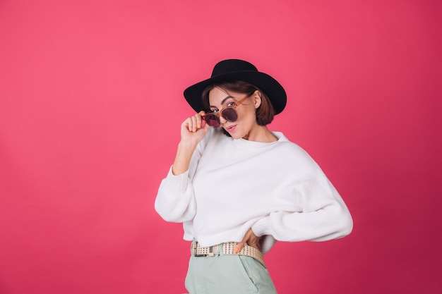 Stilvolle Frau im weißen lässigen Pullover, in der Sonnenbrille und im Hut auf der roten rosa Wand