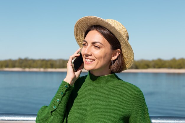 Stilvolle Frau in grünem lässigem Pullover und Hut im Freien auf Brücke mit Flussblick bei warmen sonnigen Sommertaggesprächen auf Handy