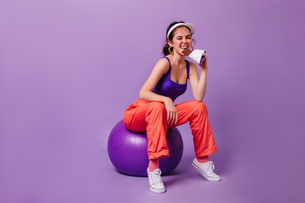 Stilvolle Frau in lila Spitze und rote Jogginghose isst Tafel Schokolade, die auf fitball gegen lila Wand sitzt