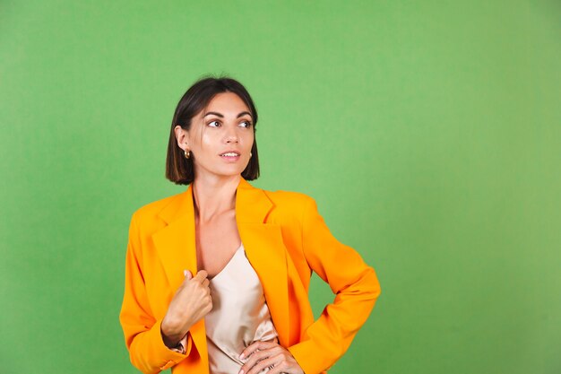 Stilvolle Frau in seidenbeigem Kleid und orangefarbenem übergroßem Blazer auf grünem, positivem Emotionenlächeln