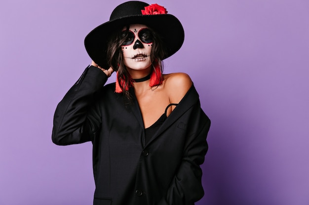 Stilvolle Frau in übergroßer Jacke und ungewöhnlicher Halloween-Maske berührt Hutkrempe. Porträt des attraktiven gebräunten Mädchens auf lila Wand.