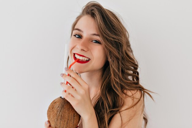 Stilvolle Frau mit roten Lippen und weißen Zähnen trinkt Kokosnuss und posiert
