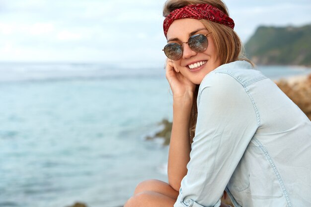 Stilvolle Frau mit Sonnenbrille, die am Strand sitzt