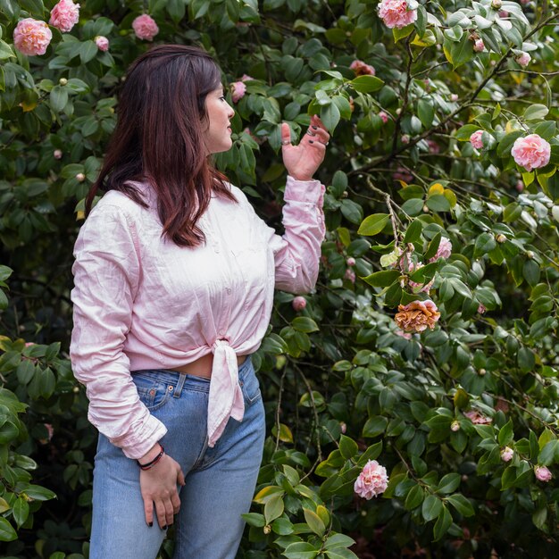 Stilvolle Frau nahe den rosa Blumen, die auf grünen Zweigen wachsen