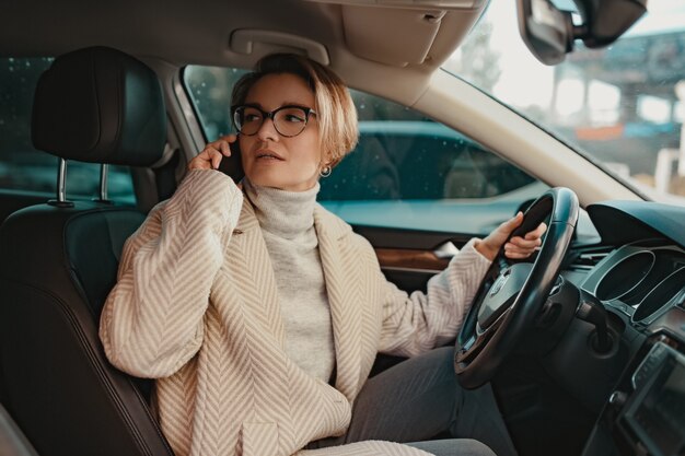 Stilvolle Frau sitzt im Auto gekleidet im Wintermantel und Brille mit Smartphone