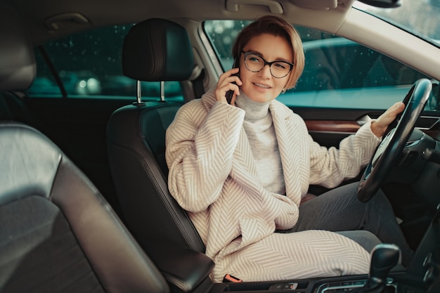 Stilvolle Frau sitzt im Auto gekleidet im Wintermantel und Brille mit Smartphone