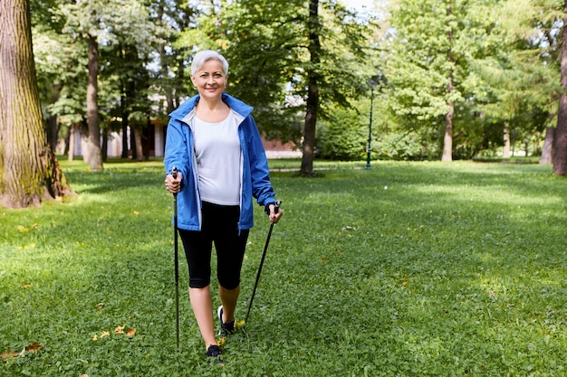 Stilvolle fröhliche ältere ältere Frau gekleidet in Sportbekleidung, die schöne wilde Natur am friedlichen Sommermorgen bewundert, mit speziellen Stöcken gehend, mit freudigem breitem Lächeln