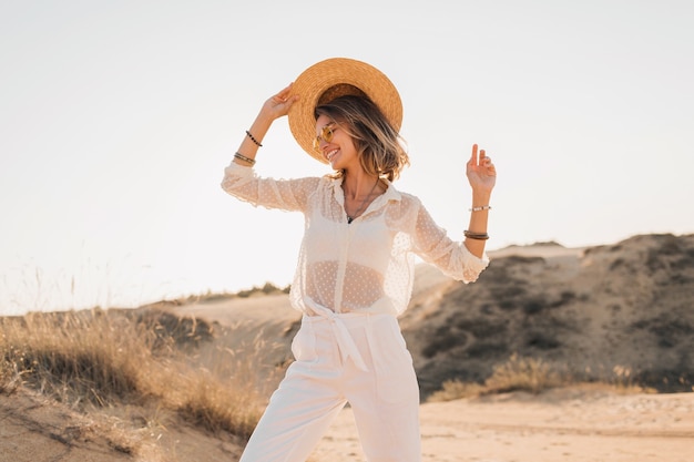 Stilvolle glückliche attraktive lächelnde Frau, die im Wüstensand posiert, gekleidet in weißen Kleidern, die Strohhut und Sonnenbrille auf Sonnenuntergang tragen