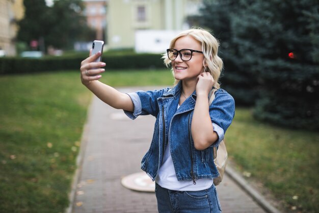 Stilvolle glückliche hübsche Mode blonde Mädchen Frau in Jeans Suite macht Selfie auf ihrem Handy in der Stadt am Morgen