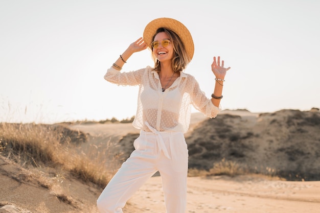 Stilvolle glückliche schöne lächelnde Frau, die im Wüstensand im weißen Outfit mit Strohhut und Sonnenbrille auf Sonnenuntergang aufwirft