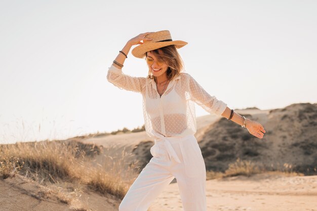 Stilvolle glückliche schöne lächelnde Frau, die im Wüstensand im weißen Outfit mit Strohhut und Sonnenbrille auf Sonnenuntergang aufwirft