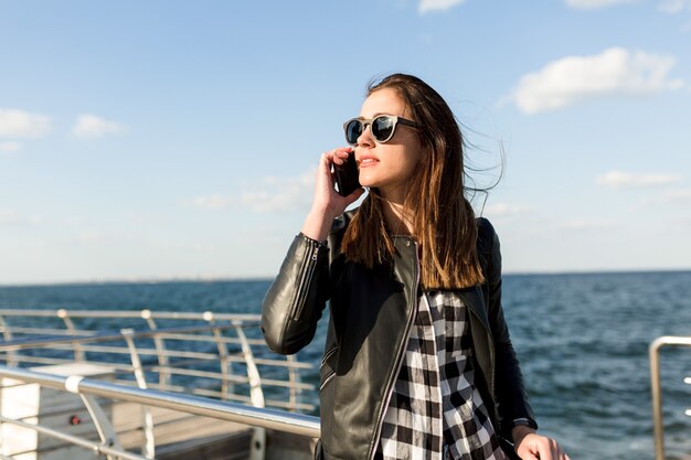 Stilvolle hübsche Frau mit dunklem Haar, das Lederjacke und Sonnenbrille trägt, die am Telefon nahe dem Meer sprechen