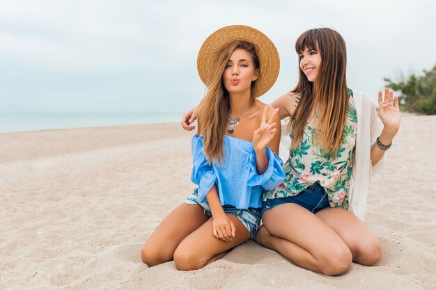 Stilvolle hübsche Frauen sitzen auf Sand in den Sommerferien am tropischen Strand, böhmischer Stil, Freunde reisen zusammen, Modetrendzubehör, lächelnde glückliche Emotion, positive Stimmung, Strohhut