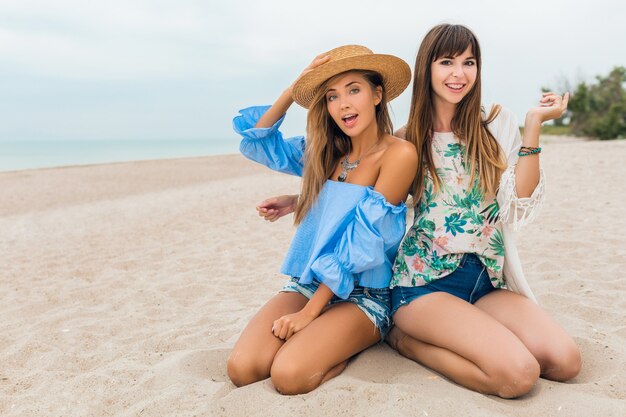 Stilvolle hübsche Frauen sitzen auf Sand in den Sommerferien am tropischen Strand, böhmischer Stil, Freunde reisen zusammen, Modetrendzubehör, lächelnde glückliche Emotion, positive Stimmung, Strohhut