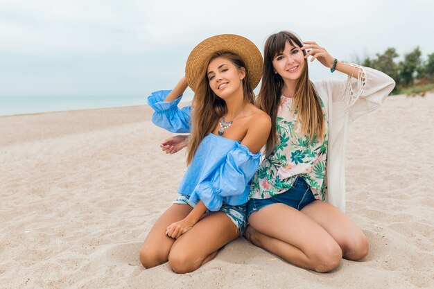 Stilvolle hübsche Frauen sitzen auf Sand in den Sommerferien am tropischen Strand, Strohhut