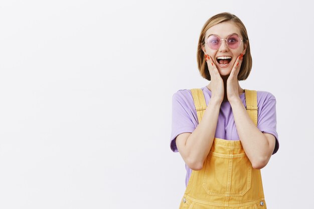 Stilvolle hübsche junge Frau in Sonnenbrille und Sommerkleidung, die sorglos aussieht, Hände faltet und aufgeregt aussieht und sich über großartige Neuigkeiten freut