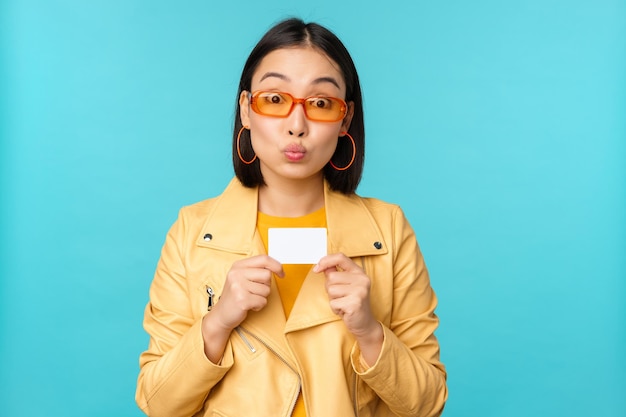 Stilvolle junge asiatische Frau mit Sonnenbrille, die Kreditkarte zeigt und lächelnd kontaktlose Bankzahlungen oder Rabatte im Laden empfiehlt, die über blauem Hintergrund stehen