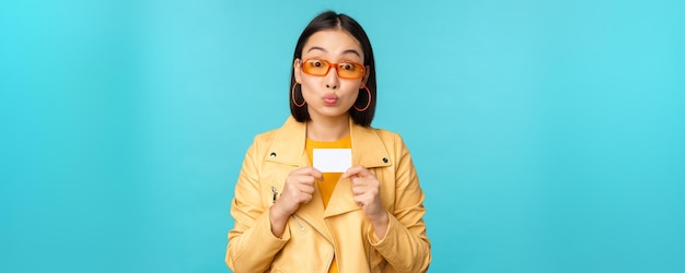 Stilvolle junge asiatische Frau mit Sonnenbrille, die Kreditkarte zeigt und lächelnd kontaktlose Bankzahlungen oder Rabatte im Laden empfiehlt, die über blauem Hintergrund stehen