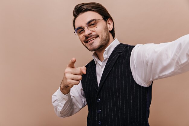 Stilvolle junge Brünette in Brille, weißem Hemd und schwarz gestreifter Weste, lächelt, zeigt und schaut in die Kamera und macht Selfie gegen eine schlichte beige Wand